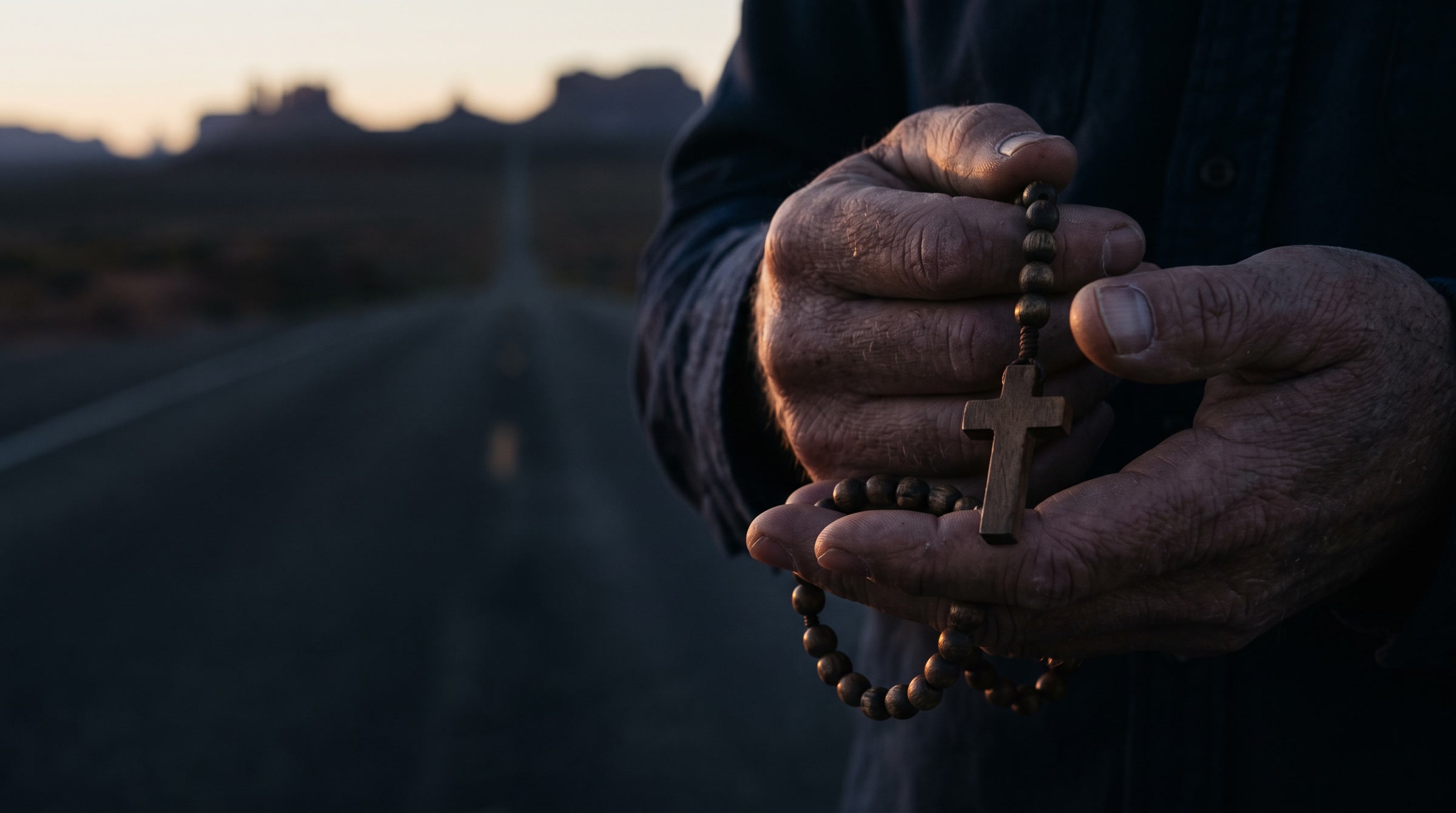 A man's weathered hands holding a wooden rosary at dusk, a desert road behind him.